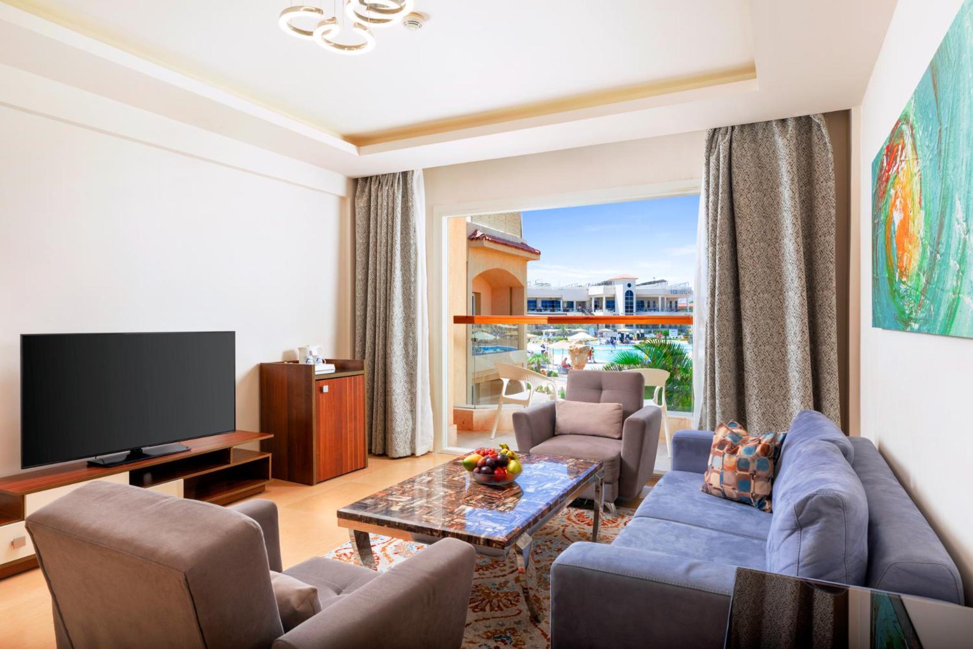 Hotel Pickalbatros Royal Moderna Sharm "Aqua Park" Exteriér fotografie The photo shows a modern and stylish living room. It features a comfortable seating arrangement with a gray sofa and two armchairs, a coffee table in the center, and a wooden TV stand with a large flat-screen TV. There are large windows with curtains