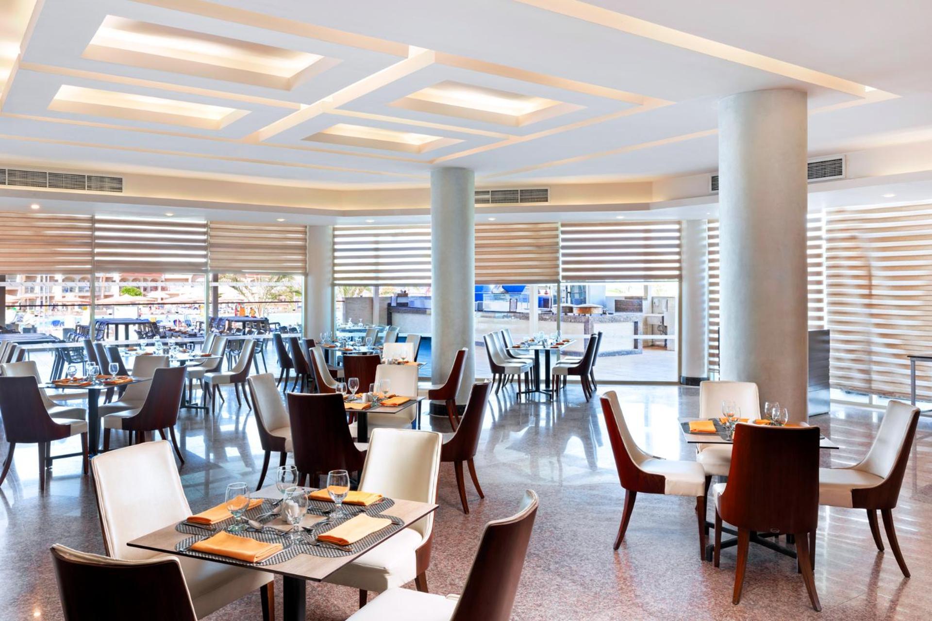 Hotel Pickalbatros Royal Moderna Sharm "Aqua Park" Exteriér fotografie The photo shows a modern restaurant interior. It features a spacious dining area with a combination of brown and white chairs arranged around tables. The design includes elegant, contemporary decor with soft lighting and a ceiling with geometric patt