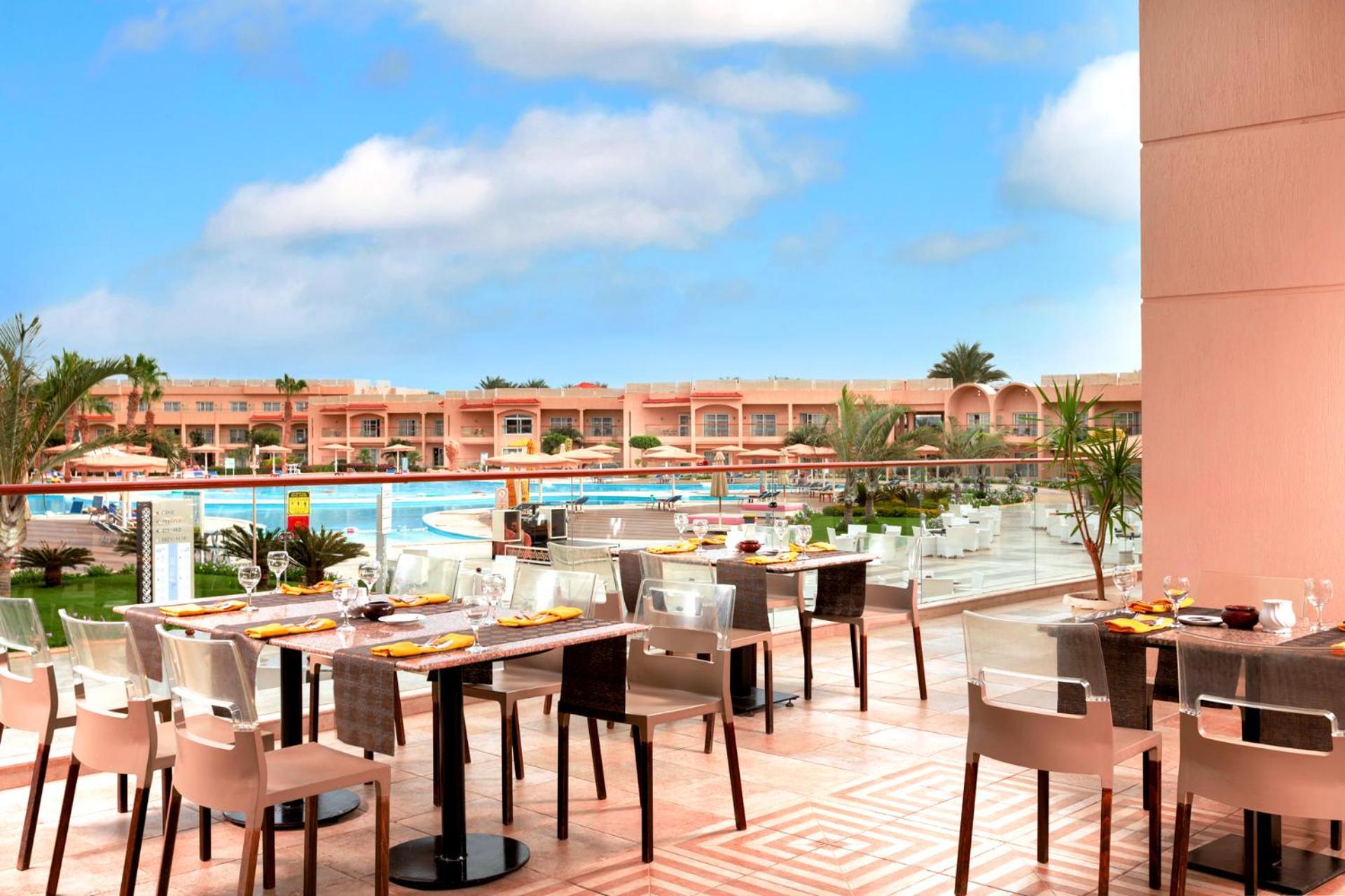 Hotel Pickalbatros Royal Moderna Sharm "Aqua Park" Exteriér fotografie The photo shows an outdoor dining area at a resort. There are several tables set for dining, featuring elegant tableware. In the background, a large swimming pool is visible, surrounded by lush greenery and lounge chairs. The buildings beyond the poo