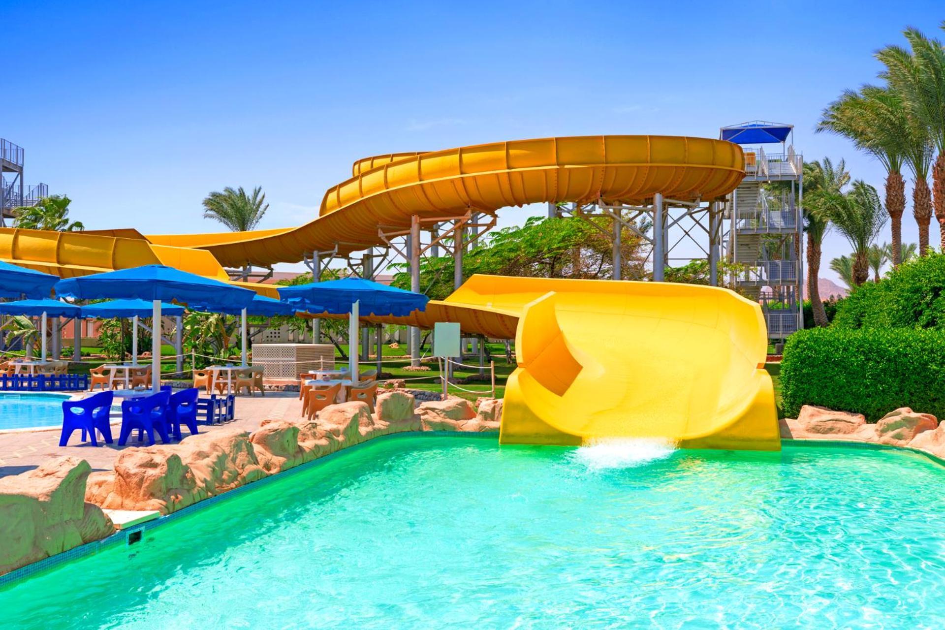 Hotel Pickalbatros Royal Moderna Sharm "Aqua Park" Exteriér fotografie The photo depicts a vibrant water park scene featuring a large yellow water slide that curves down into a clear blue swimming pool. Surrounding the slide are several blue umbrellas and seating areas, likely for guests to relax. The backdrop includes 