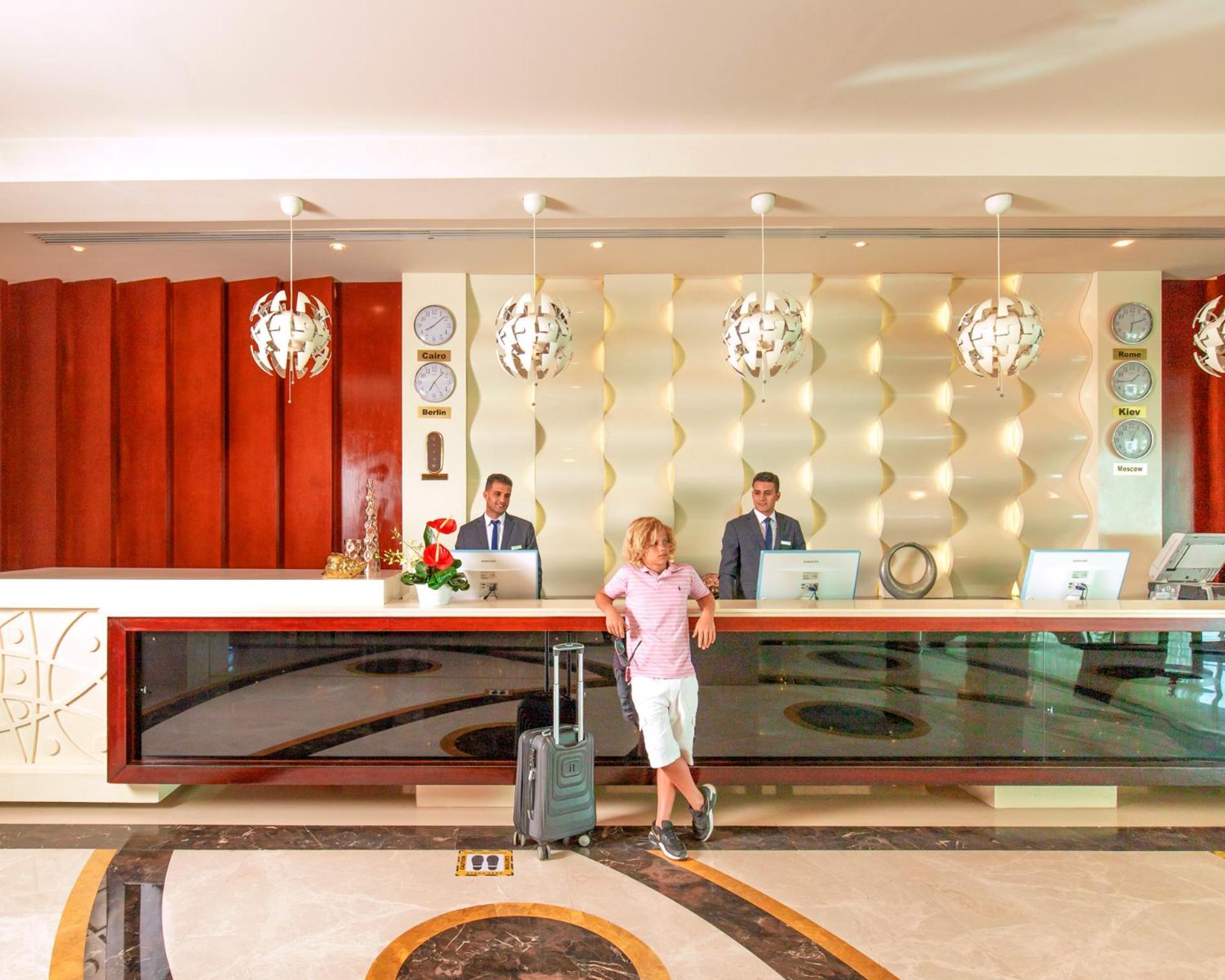 Hotel Pickalbatros Royal Moderna Sharm "Aqua Park" Exteriér fotografie The photo depicts a hotel reception area. At the front desk, two staff members in suits are standing, presumably ready to assist guests. A man in casual attire, holding a suitcase, is standing in front of the desk. The reception has a modern design w