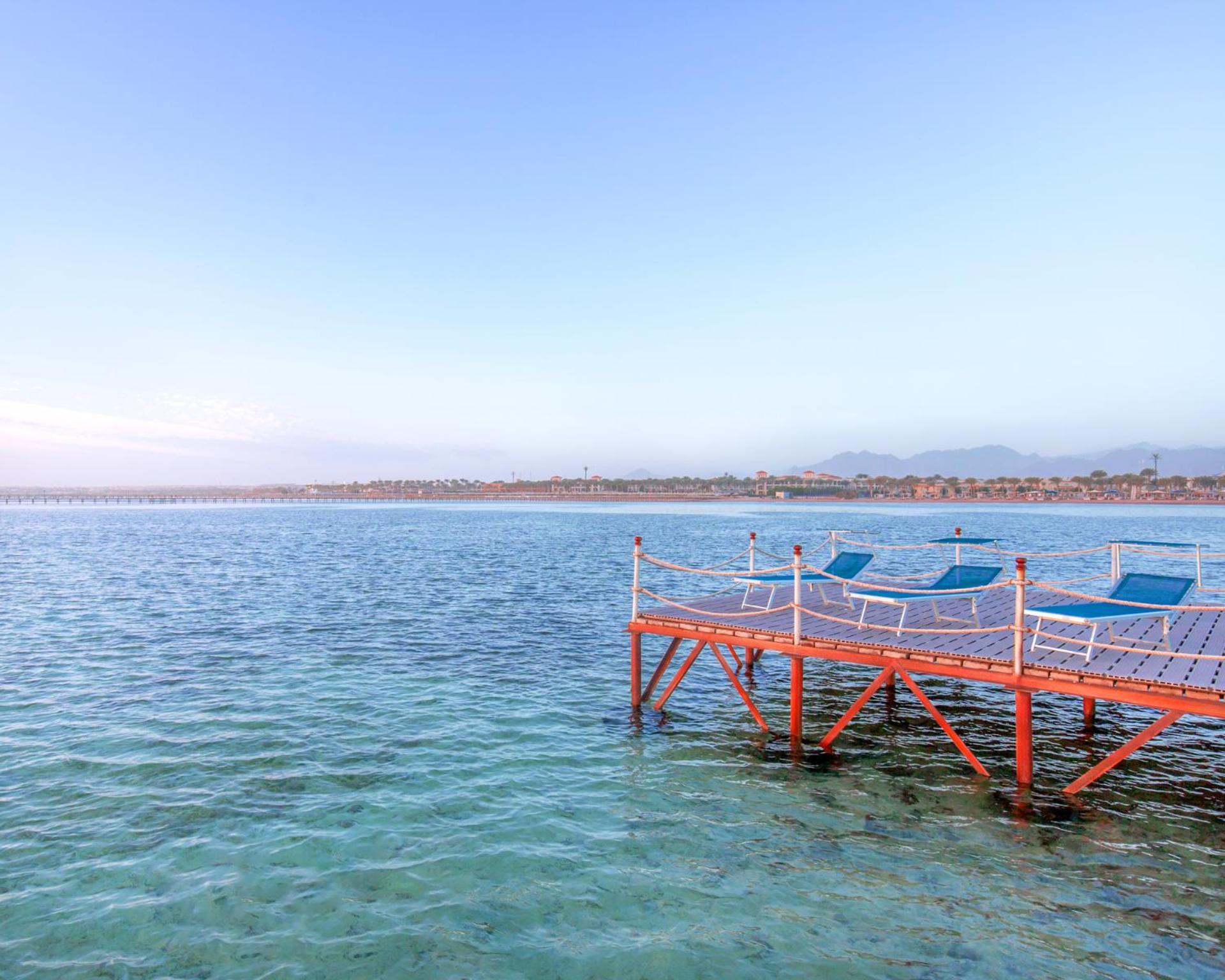 Hotel Pickalbatros Royal Moderna Sharm "Aqua Park" Exteriér fotografie The photo shows a peaceful scene featuring a wooden dock extending into calm water. The dock is painted orange and has several blue lounge chairs placed on it, suggesting a relaxed atmosphere. In the background, there's a shoreline with some greenery