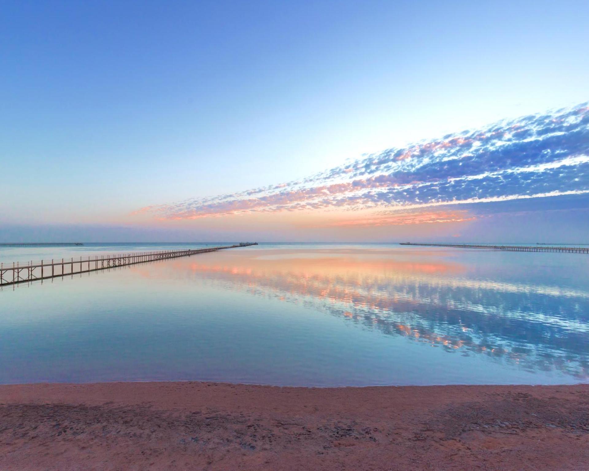 Hotel Pickalbatros Royal Moderna Sharm "Aqua Park" Exteriér fotografie The photo depicts a serene coastal scene at dawn or dusk. There is a calm body of water that reflects the soft hues of the sky, which include shades of blue, pink, and orange. A long wooden pier extends into the water, adding depth to the composition